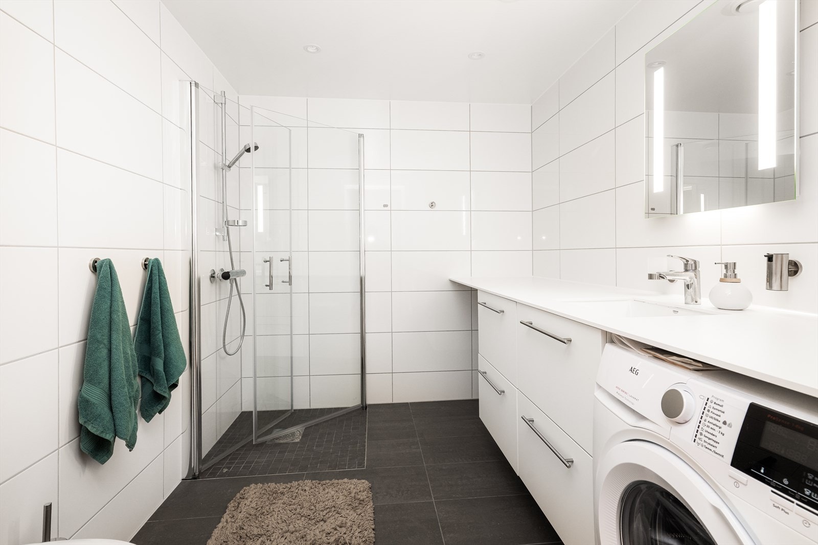 Under benken er det innfelt vaskemaskin samt gode skuffer og åpen plass til vaskekurv. Badet har speil med integrert belysning samt down-light belysning i tak. I tillegg har badet dusjhjørne med innfellbare dører i herdet glass og veggmontert toalett. Galleribilde