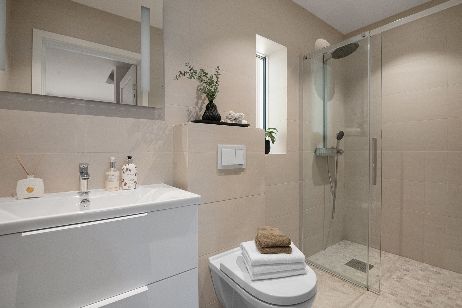 Badet i 1. etasje fremstår moderne og tidløst, med helflisede flater i rolige toner. Dusjhjørne med glass, vegghengt toalett og servantskap gir et stramt og elegant uttrykk. Varmekabler i gulvet og balansert ventilasjon gir høy komfort. Galleribilde