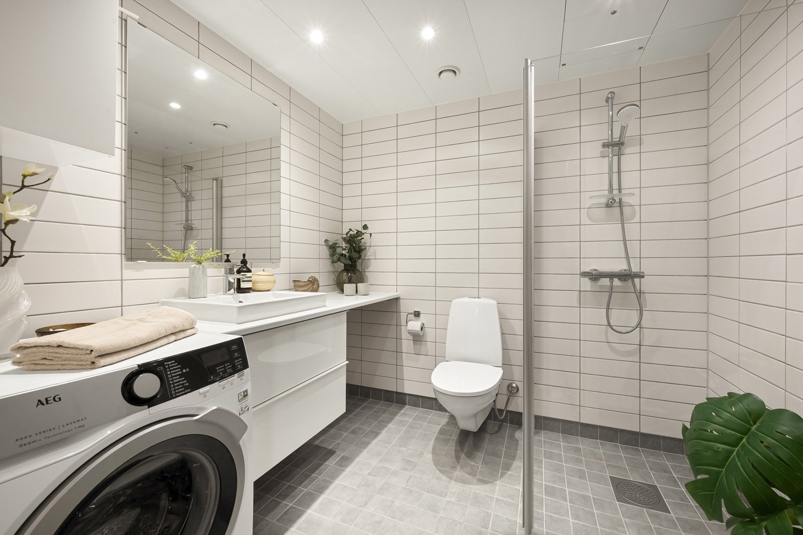 Badet er innredet med vegghengt toalett, dusjhjørne med svingbare glassdører og bred baderomsinnredning med skuffer, nedfelt servant, benkeplate og vegghengt speil. Galleribilde