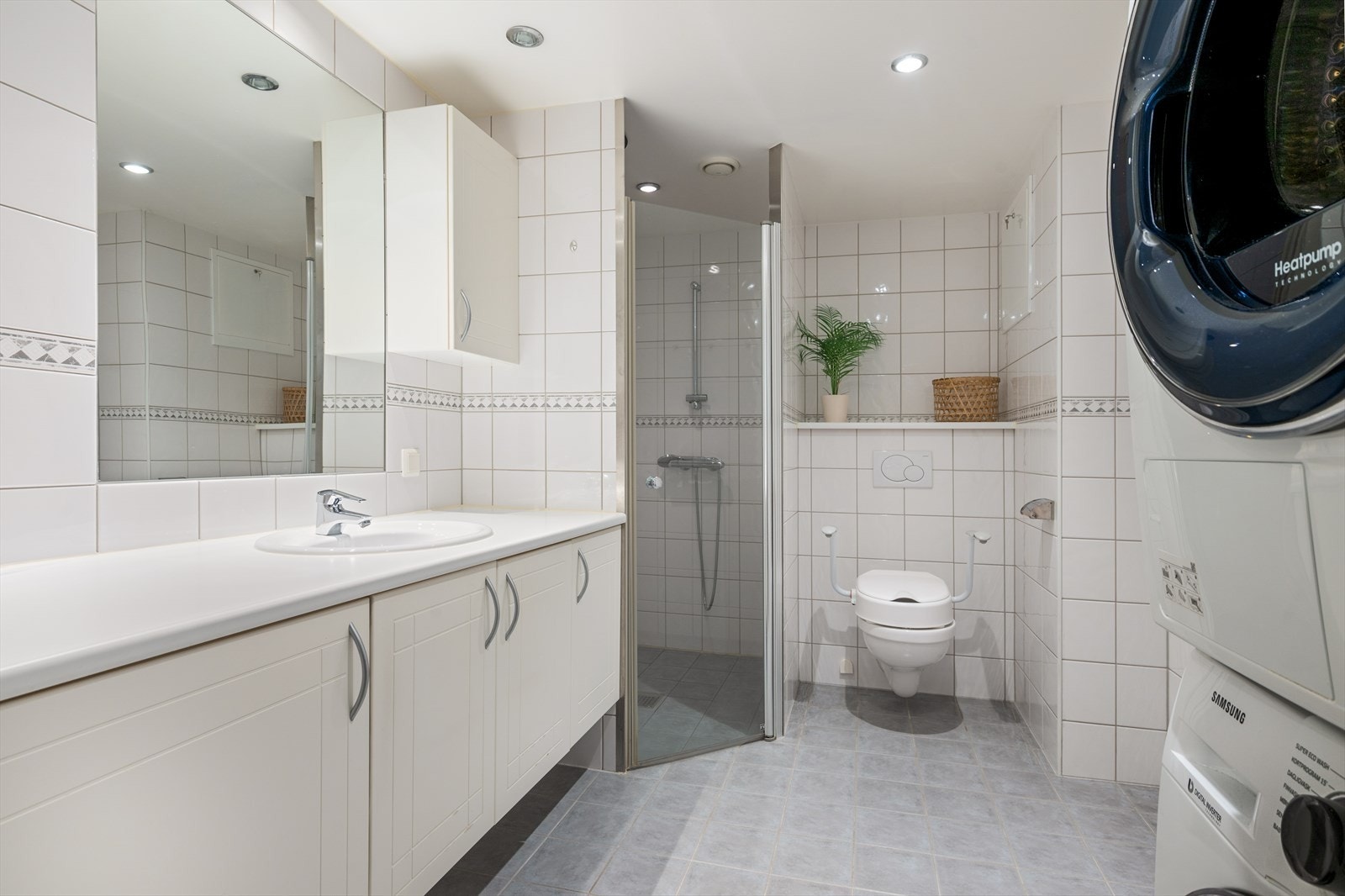 Flislagt baderom med tidløse fliser, dusjnisje, vegghengt wc og nedfelt servant på baderomsinnredning. Det er plass til vaskemaskin og tørketrommel i vaskesøyle. Galleribilde