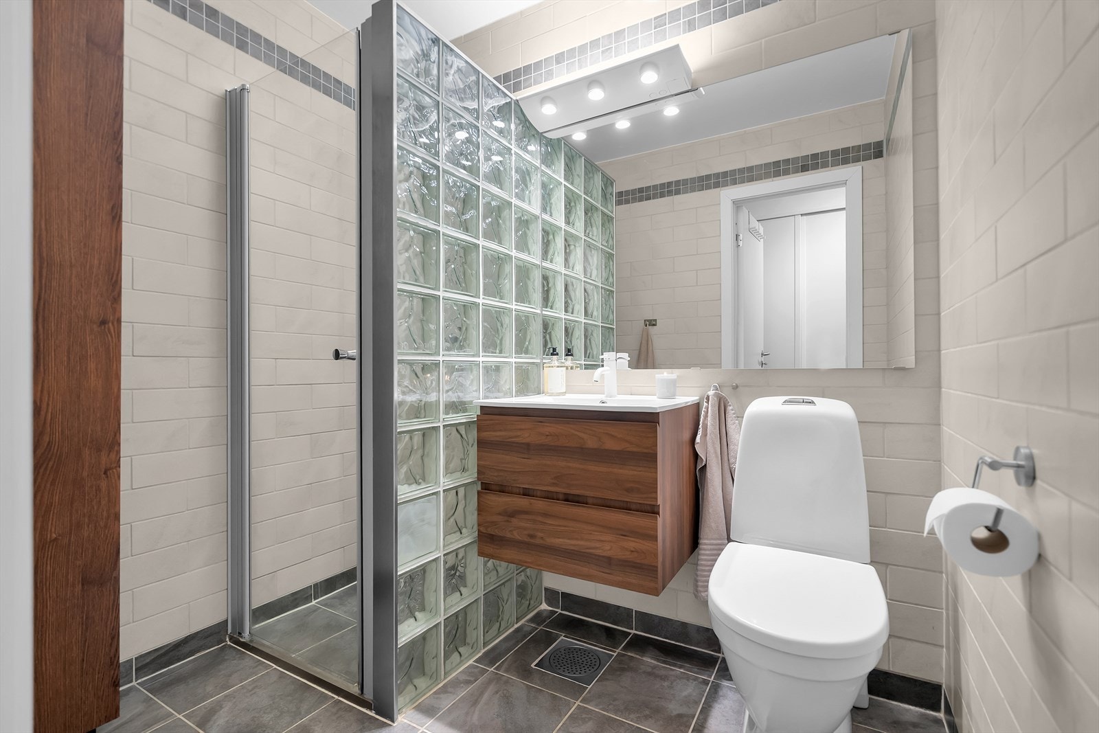 Pent helfliset baderom som fremstår som moderne og funksjonelt. Badet har en dusjnisje med glassdør, servant med underskap og toalett. Gulvet er utstyrt med varmekabler for ekstra komfort, og mekanisk avtrekk sikrer god ventilasjon. Galleribilde