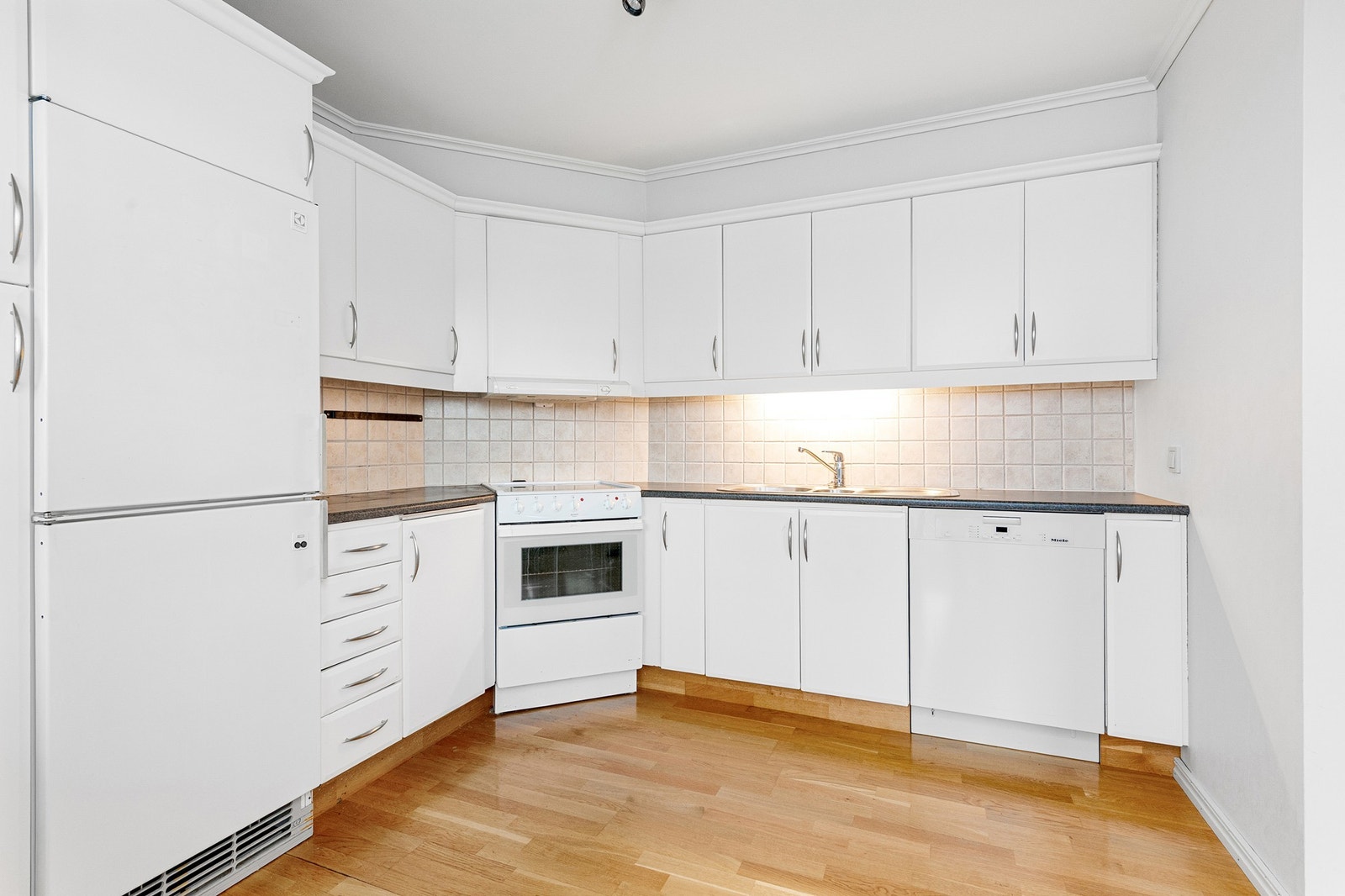 Kjøkkeninnredning med hvite fronter. Laminat benkeplate. Det er fliser mellom benken og overskapene. Innredningen er utstyrt med komfyr og med opplegg for oppvaskmaskin. Det er avtrekk via balansert eller mekanisk ventilasjon anlegg Galleribilde