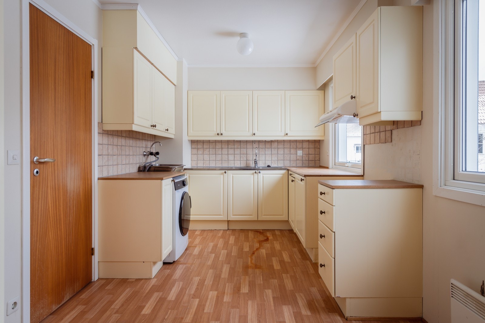 Kjøkkeninnredning med malte speilfronter, laminat benkeplate, stål oppvaskkum og frittstående hvitevarer. Det er opplegg for vaskemaskin i rommet. Galleribilde