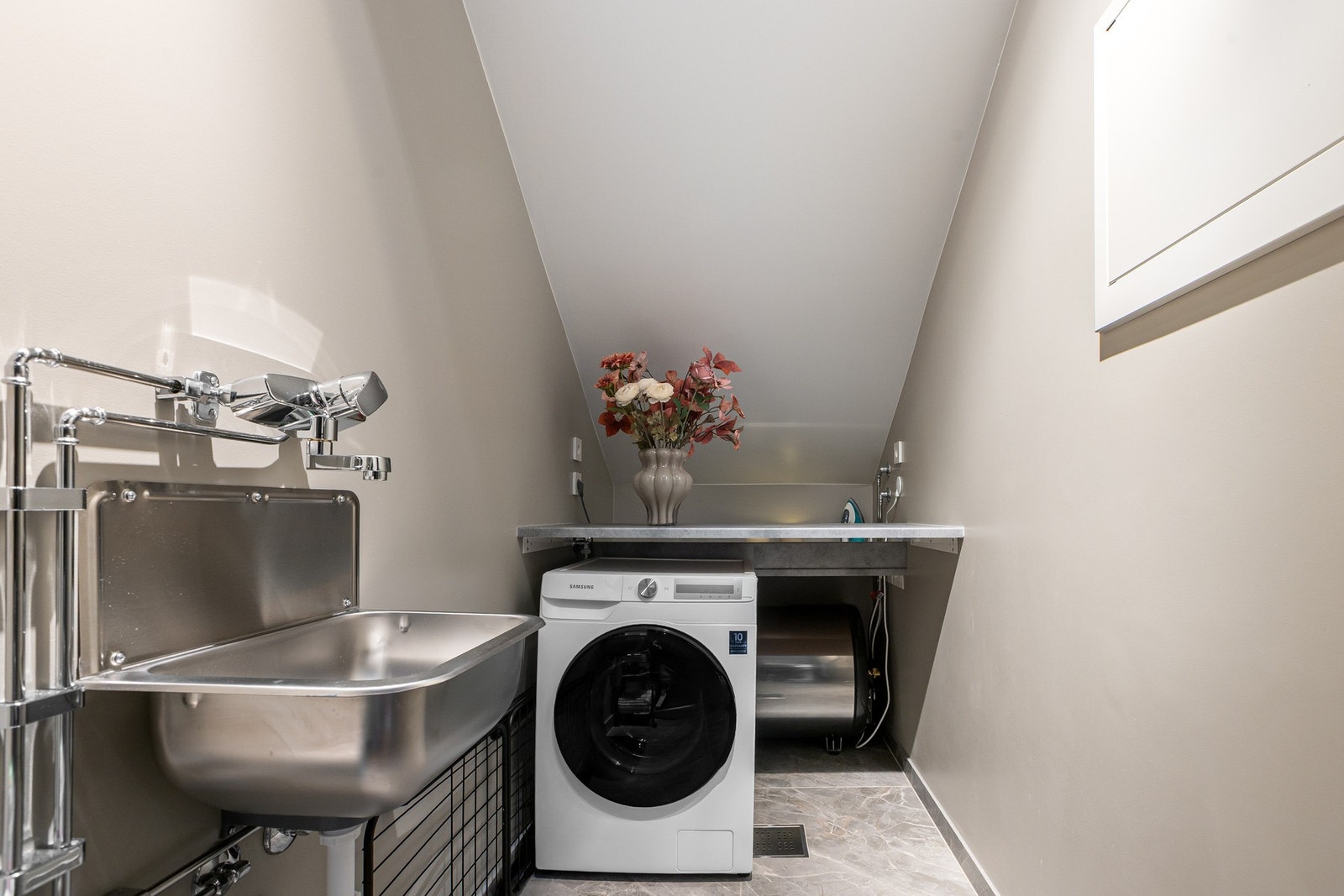 Praktisk vaskerom med flislagt gulv og gulvvarme, utslagsvask samt nisje for vaskemaskin og tørketrommel under benkeplate. Galleribilde