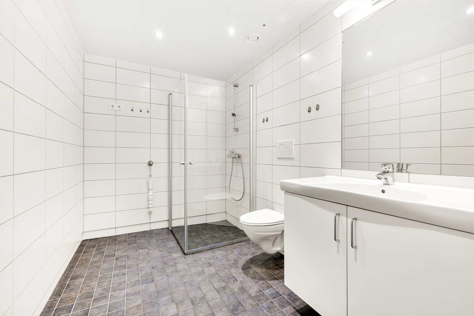 Delikat, flislagt bad med vegghengt toalett og dusjhjørne med innfellbare glassdører. Badet har behagelige varmekabler i gulvet og praktisk opplegg for vaskemaskin. Galleribilde