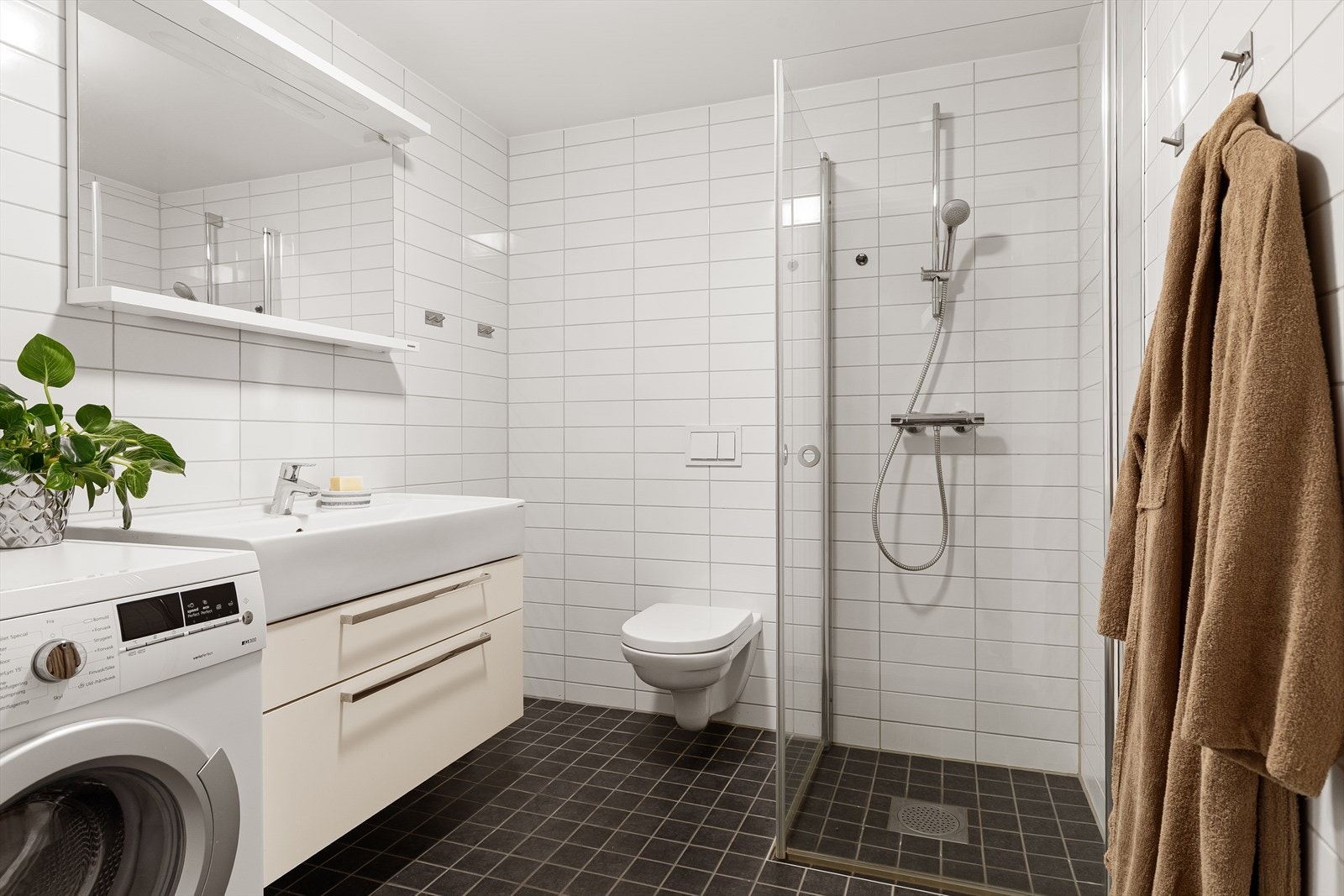 Leiligheten har et kombinert bad/vaskerom som er lyst og funksjonelt innredet med servantskap med nedfelt servant, speil med belysning, vegghengt klosett med innebygd sisterne og dusjsone med innfelte dusjdører i herdet glass. Galleribilde