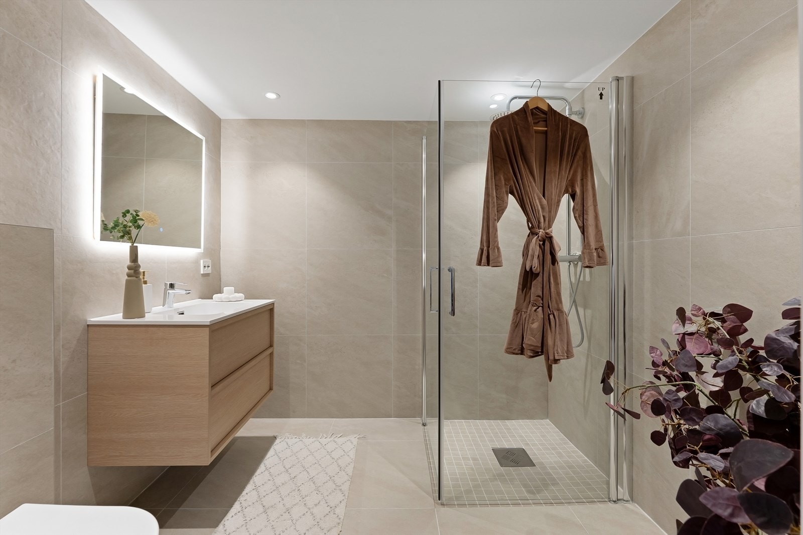 Bad fra 2025 med dusjhjørne med dører i herdet glass, speil med LED lys over servant, vegghengt toalett, flislagt gulv med varmekabler og elektrisk styrt vifte. Galleribilde