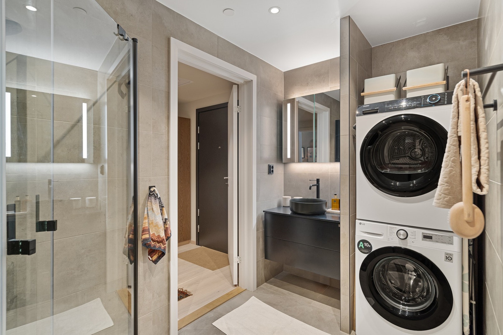 Speil med integrert belysning og downlights i himling gir behagelig lys. Badet er også praktisk innredet med søyle for vaskemaskin og tørketrommel, diskret plassert og lett tilgjengelig fra gangen. Dette gjør rommet funksjonelt som kombinert bad/vaskerom. Galleribilde