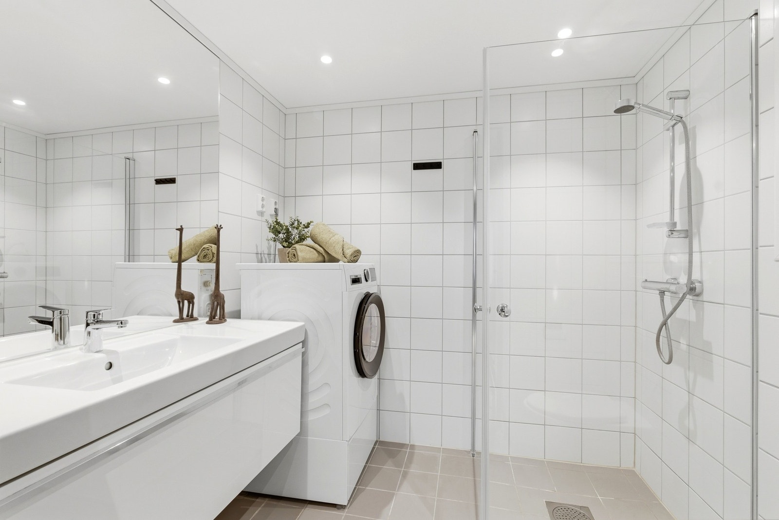 Stilrent bad med lyse vegg- og gulvfliser. Badet har moderne innredning, vegghengt wc, dusjhjørne og opplegg for vaskemaskin. Galleribilde