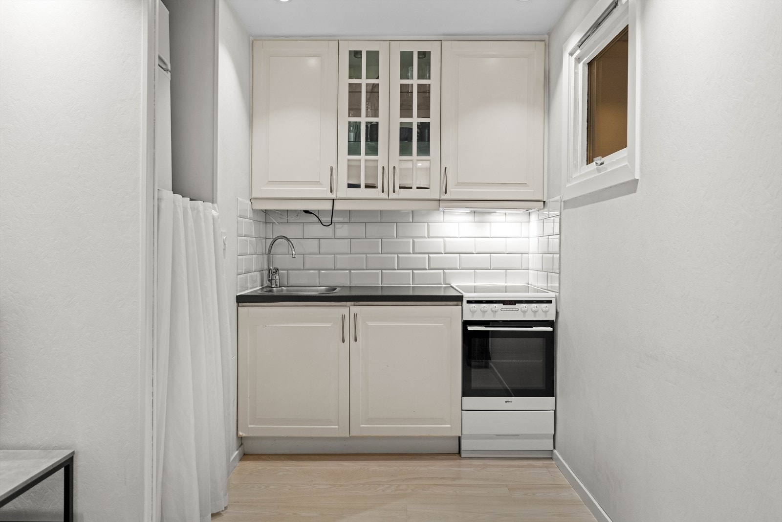 Kjøkkeninnredning med profilerte fronter og enkelte overskap med glassfronter. Fliser på vegg over benkeskap, laminat benkeplate med nedfelt oppvaskkum i rustfritt stål samt ventilator over kokesone. Galleribilde