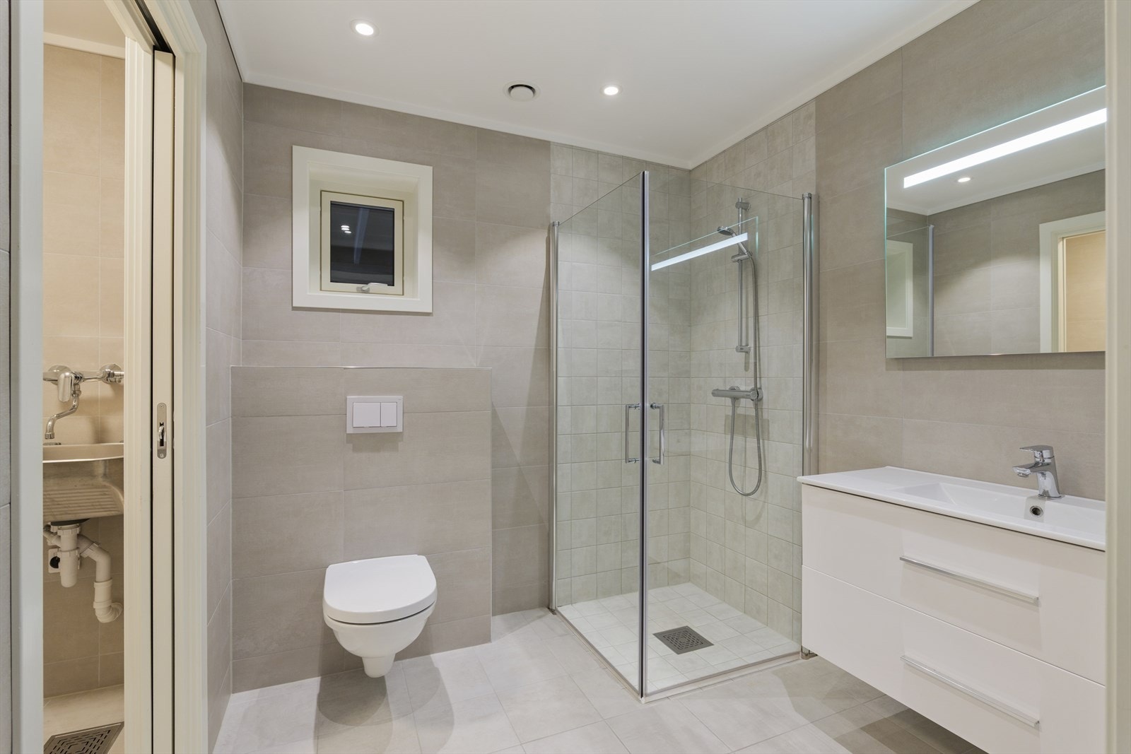 Baderommet er utstyrt med baderomsinnredning med servant, vegghengt toalett og dusjhjørne. Ventilering via via balansert ventilasjon (avtrekksventil i tak samt luftespalte i dør). Galleribilde