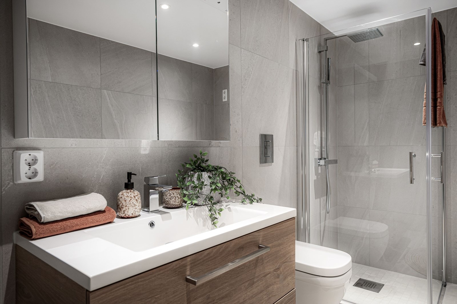 Baderomsinnredningen fra Bano Home med hvit heldekkende servant, speilskap med lys i toppen av dørene, vegghengt toalett med softclose, spyleknapp fra Blank krom, dusjvegger, servantarmatur fra Grohe Eurocube og dusjsett fra Grohe Eurocube. Galleribilde