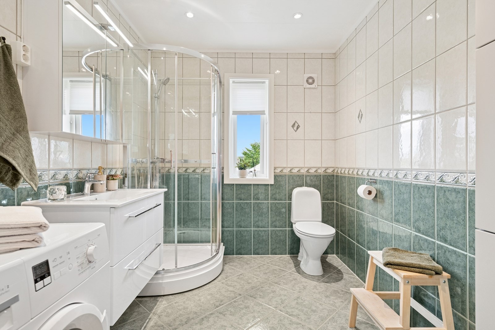 Bad som inneholder dusjhjørne, toalett, servant med over- og underskap og opplegg til vaskemaskin Galleribilde