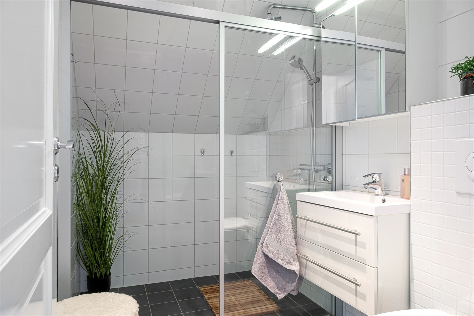 Badet fremstår som funksjonelt, tidløst, med lyse veggfliser som står i kontrast til mørke gulvfliser. Det er vegghengt toalett, dusj, servant med underskap og overhengt speil med skap. Galleribilde