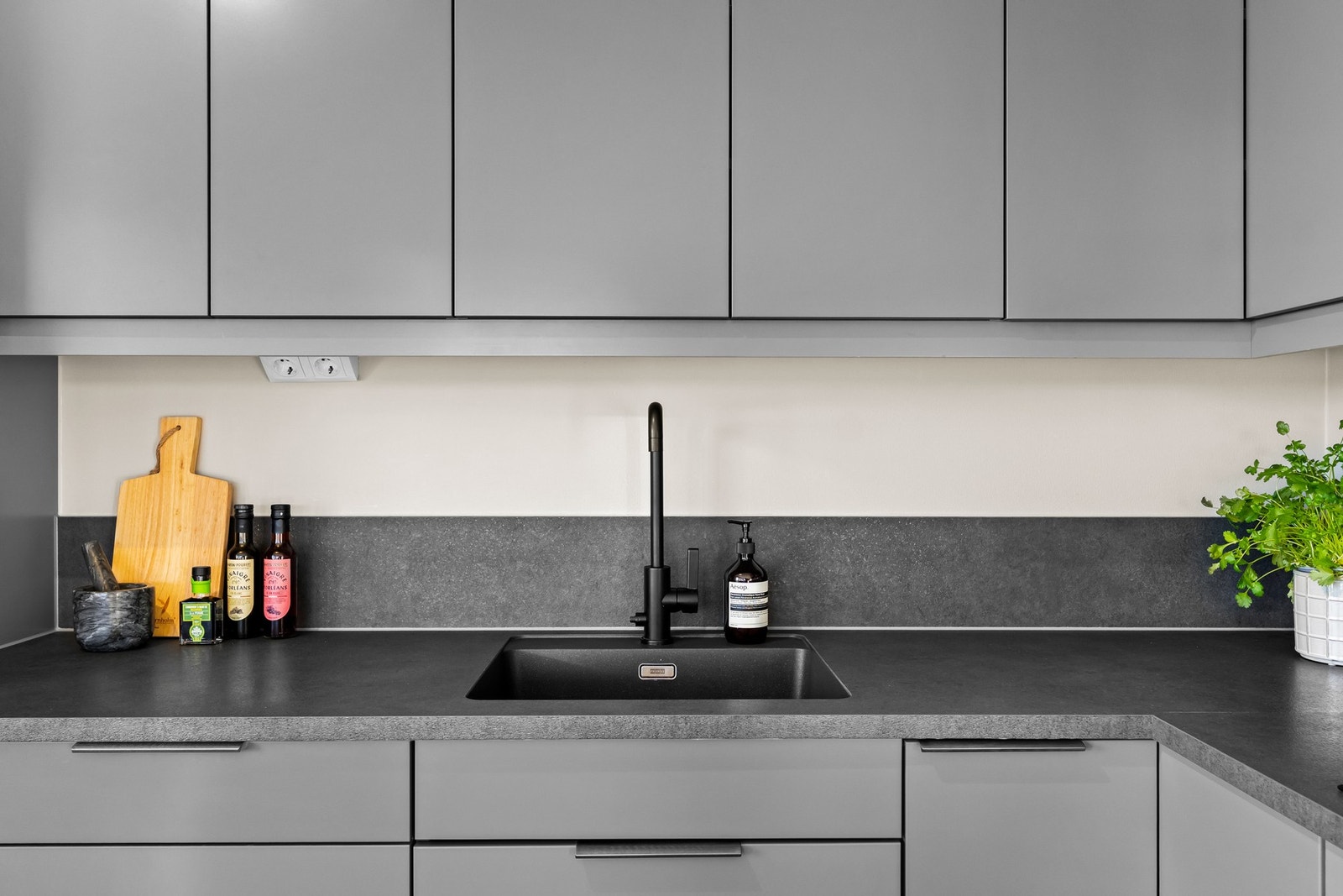 Glatte grå fronter, gråsort, laminat benkeplate med sort planlimt kim i kompositt, sort blandebatteri fra Tapwell, og backsplash benkeplate på vegg. Galleribilde