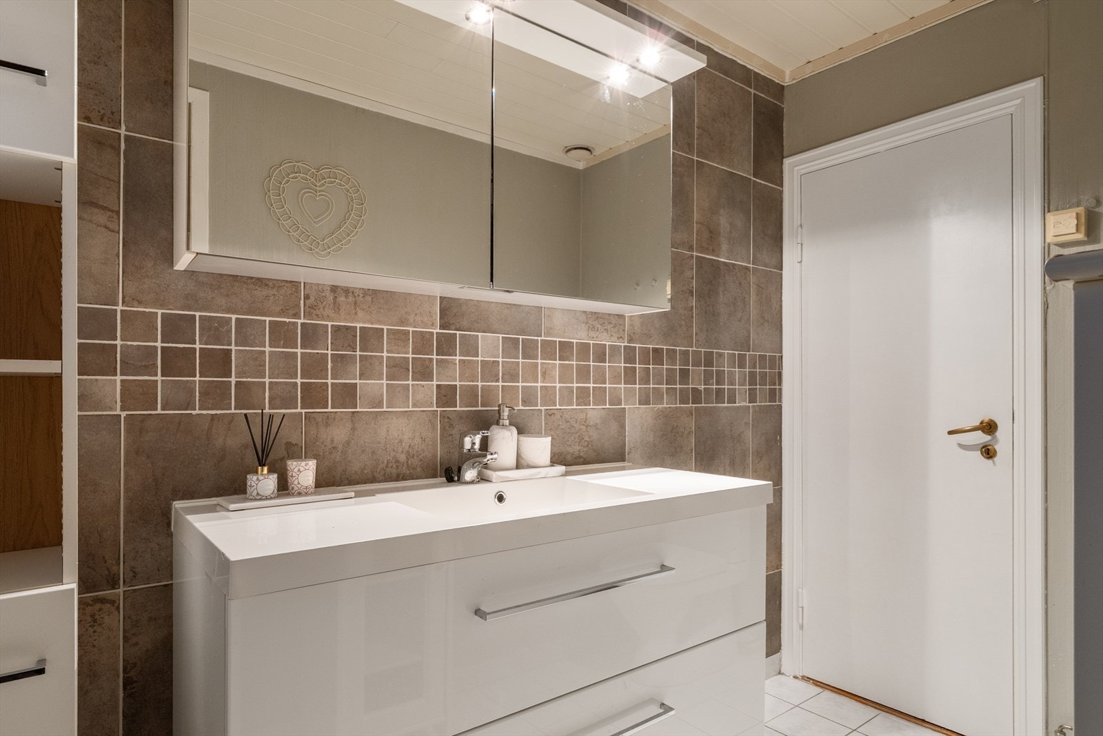 Badet er innredet med et servantskap med servant, tilhørende speilskap og høyskap, et gulvmontert toalett og en vegghengt dusj. Galleribilde