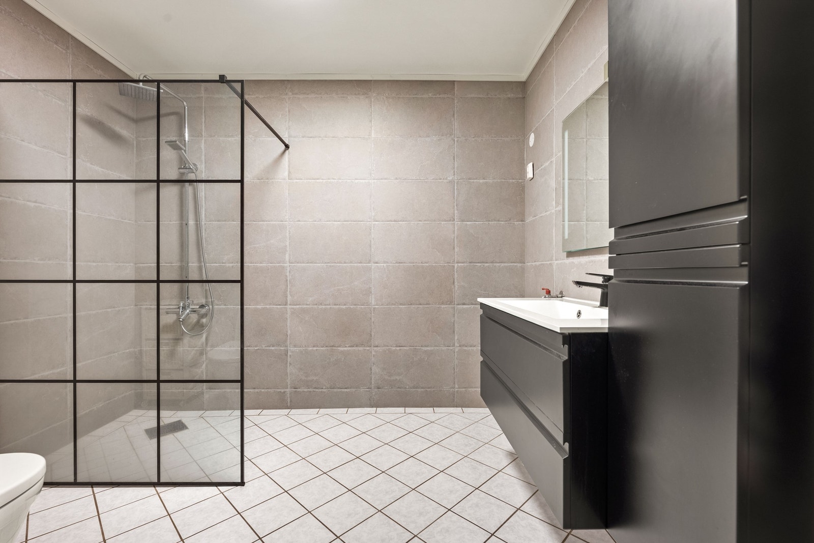 Badet har dusj i hjørne med dusjvegg av glass samt veggmontert regnfallsdusj Galleribilde