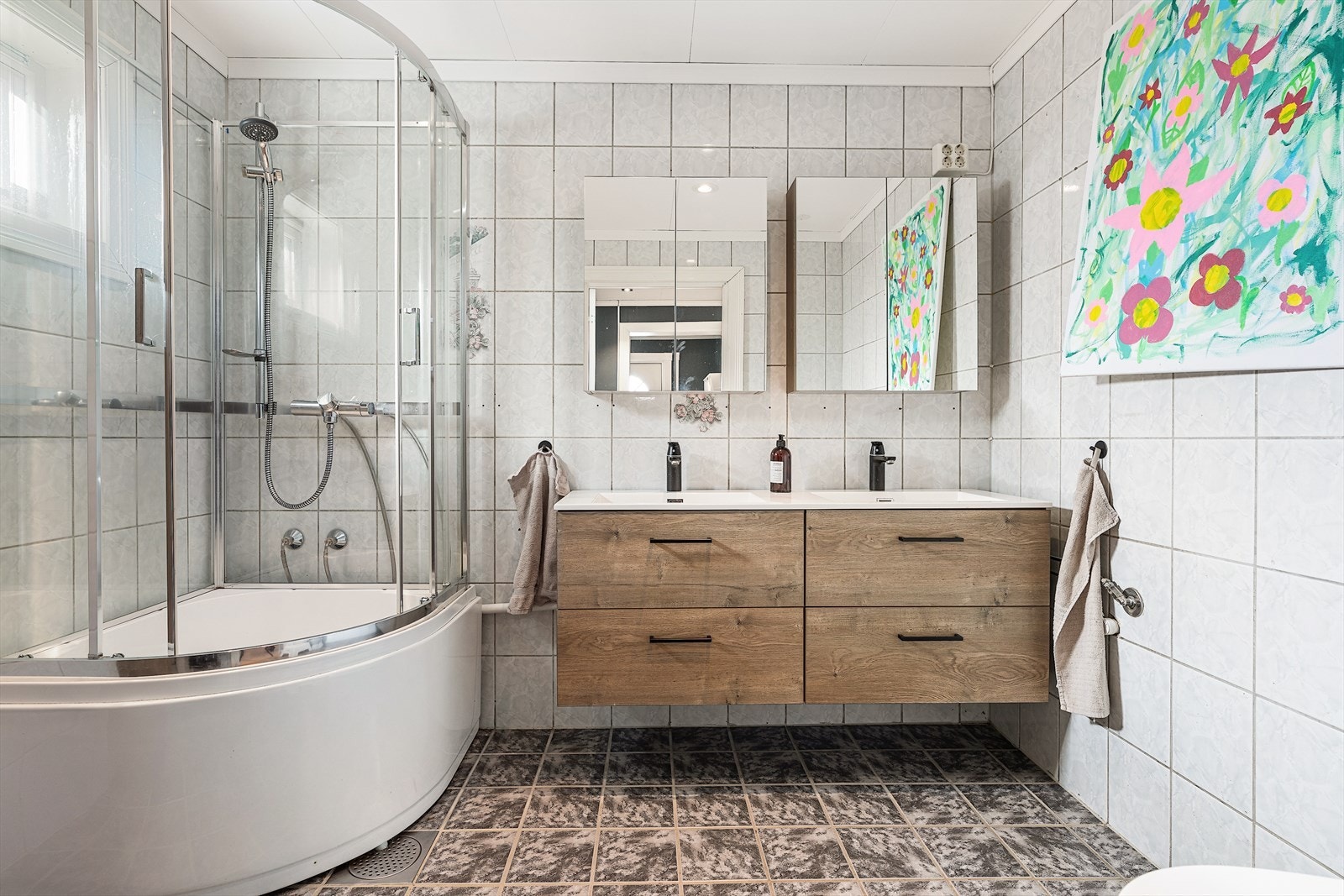 Lyst og romslig baderom med pene flislagte overflater. Baderomsinnredningen består av toalett, dobbelservant med underskuffer, to speilskap, og badekar med dusj og kabinett Galleribilde