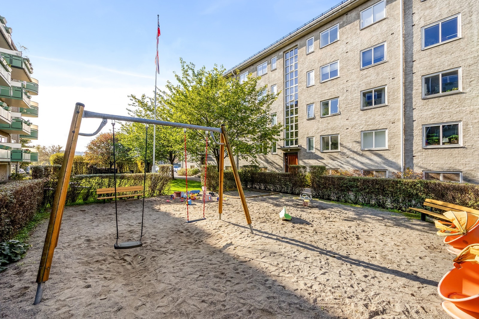 Hyggelig lekeplass i gårdsrommet med husker, sandkasse og benker omgitt av grønt. Galleribilde