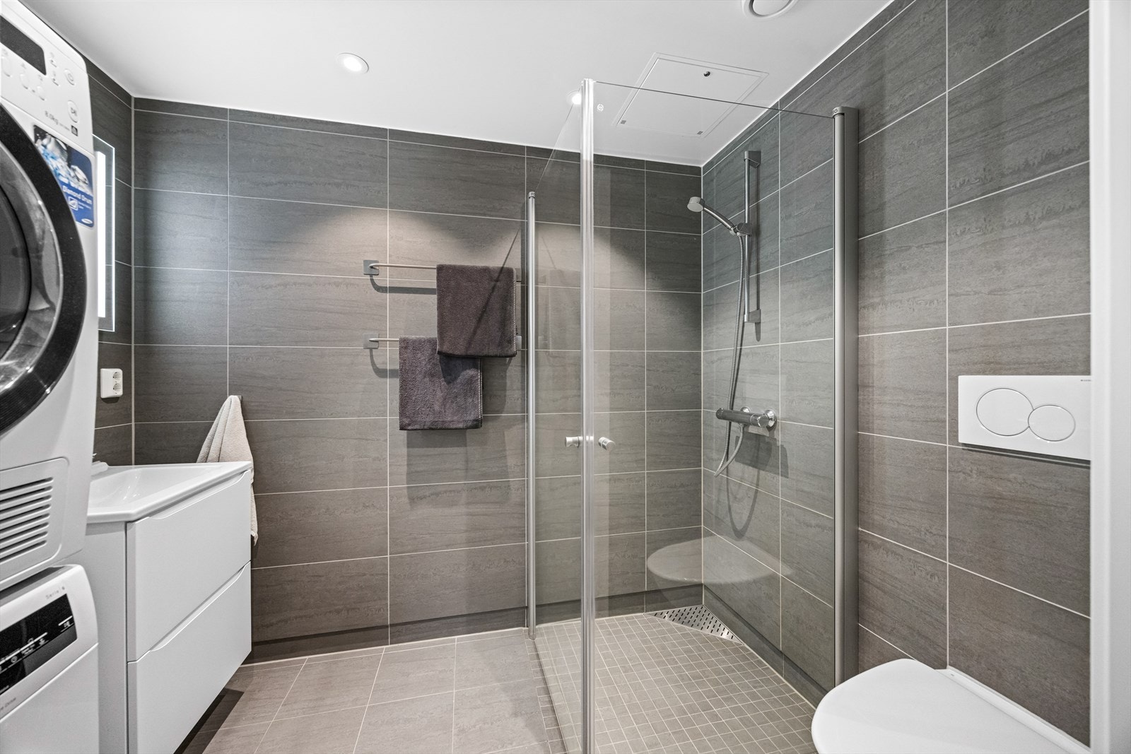 Badet er stilrent innredet med en heldekkende skuffeservant med håndtaksfrie skuffer, et moderne speil med integrert belysning, et dusjhjørne med innfellbare glassvegger, samt et moderne vegghengt toalett med skjult sisterne. Galleribilde