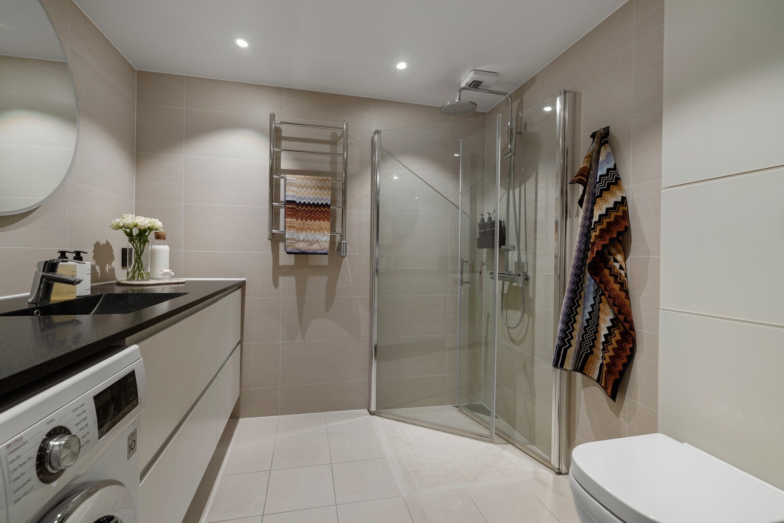 Badet har dusjhjørne med glassdører, vegghengt toalett samt baderomsinnredning med nedsunket servant og speil med lys. Det er også opplegg for vaskemaskin, noe som gir en praktisk og funksjonell løsning. Galleribilde