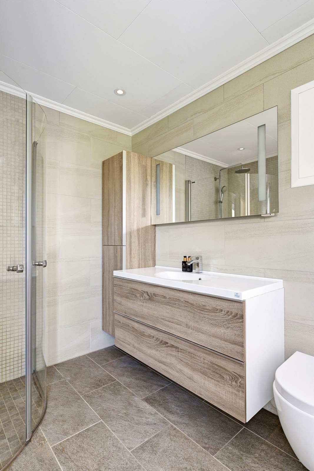 Videre er badet innredet med vegghengt toalett og innredning med heldekkende servant, speil og speilskap, dusjnisje med glassdør. Galleribilde