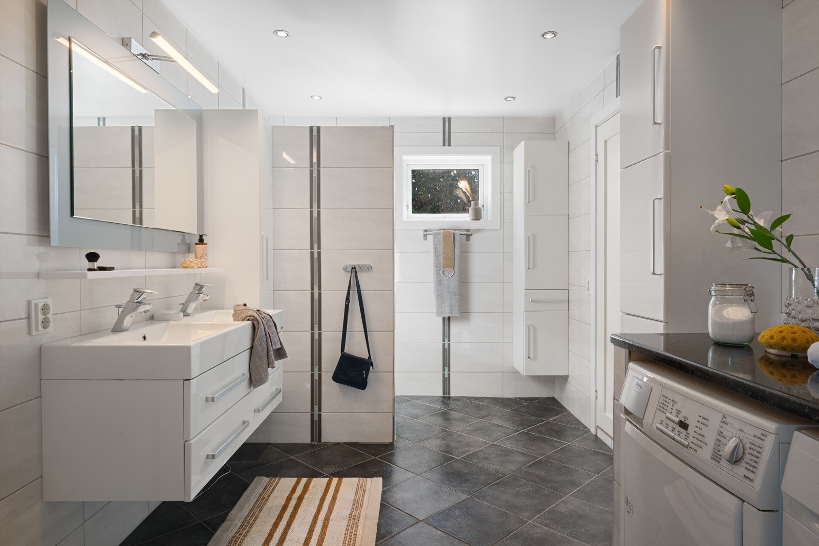 Romslig helfliset baderom med tidløs innredning. Her er det vegghengt toalett, heldekkende dobbel servant med belyst speil over, dusj og baderomsmøblement. Galleribilde