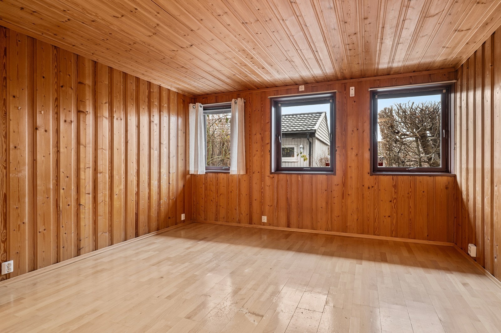 Stuen/oppholdsrommet i hybelen har vegger og himling kledd med trepanel. Tre vinduer slipper inn godt med dagslys. Galleribilde