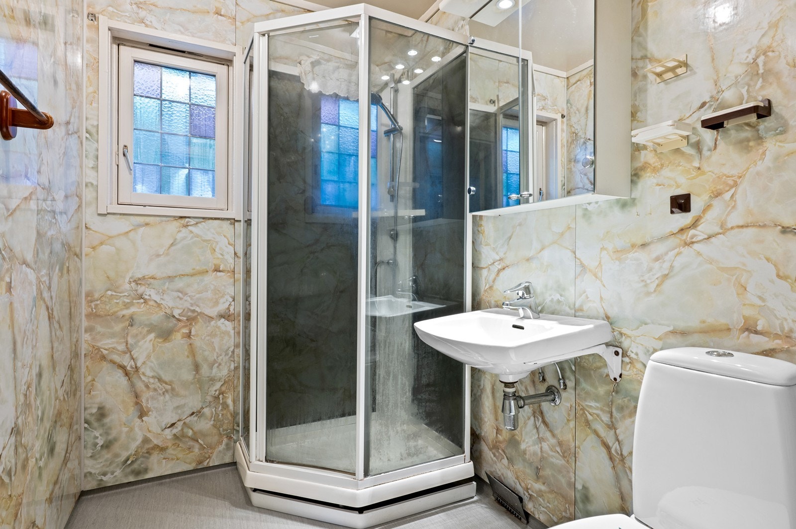 Badet i første etasje har en eldre standard, med belegg med oppbrett langs veggene. På veggene er det våtromsplater, og i himlingen er det malte plater. Rommet er innredet med dusjkabinett, servant med overskap og gulvmontert wc. Rommet bør oppgraderes. Galleribilde