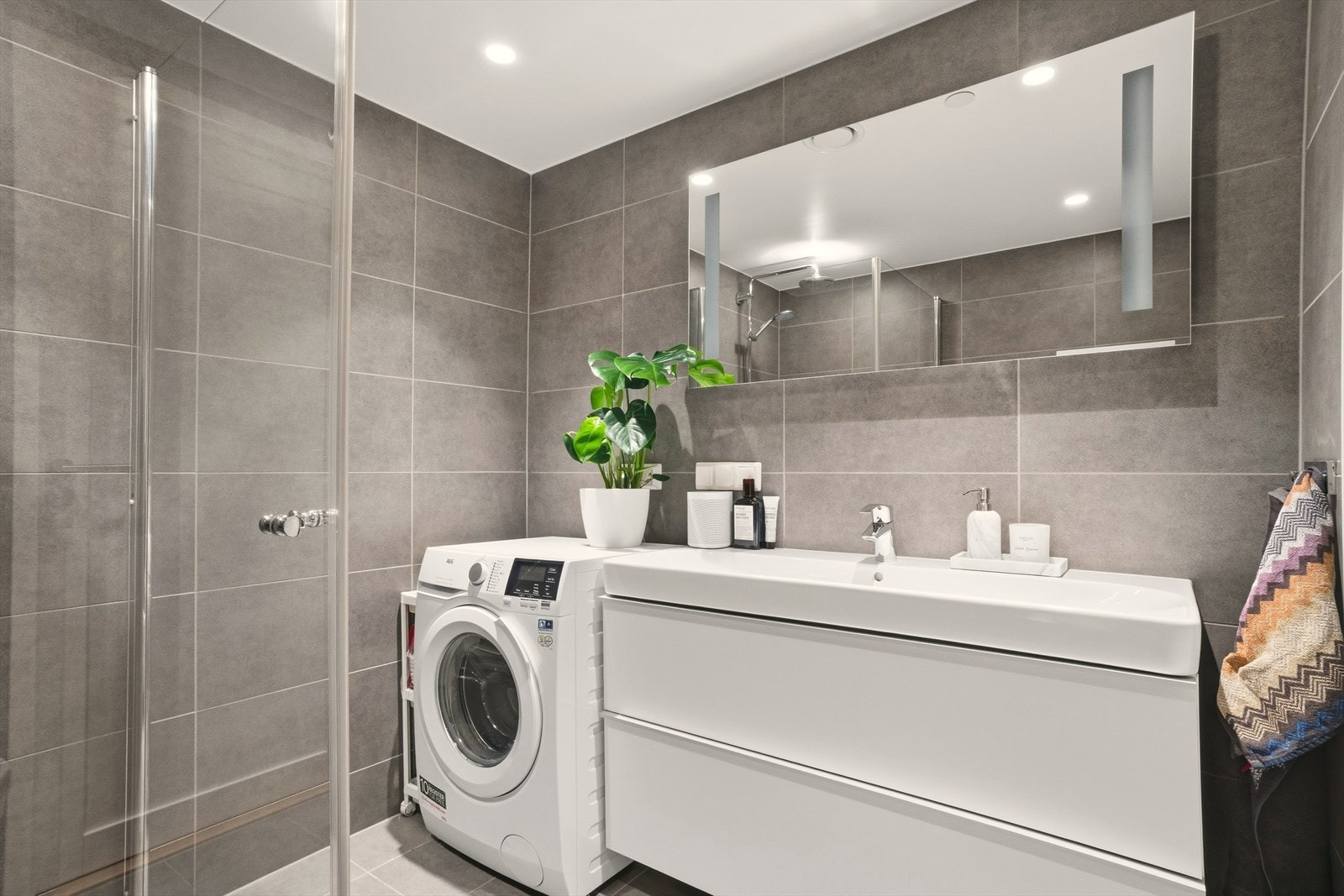 Badet inneholder innredning med heldekkende servant, speil med innfelt lys, veggmontert toalett og dusjhjørne med glassdører. Det er også opplegg og plass til vaskemaskin og tørketrommel på badet. Galleribilde