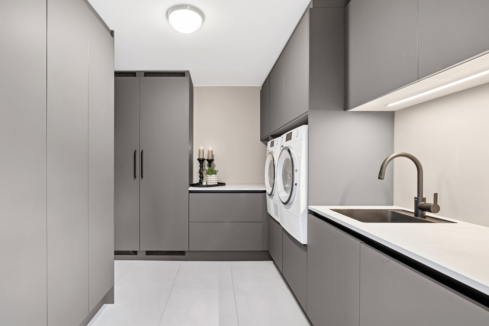 Praktisk vaskerom i 1. etasje med slettmalte vegger og fliser på gulv med gulvvarme. Innredet med diverse skap, benkeplate med vaskekum og opplegg for vaskemaskin. Galleribilde