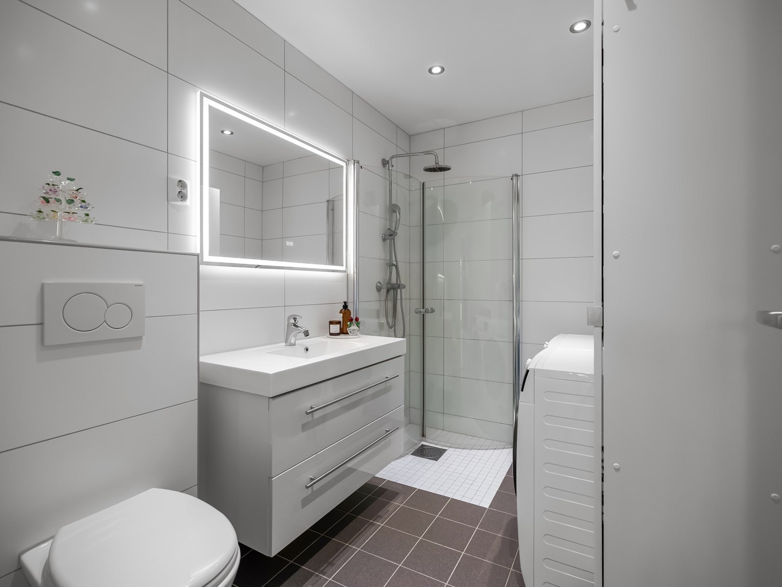 Vi er nå på badet i underetasjen, som er utstyrt med toalett, servant med oppbevaring i underskap, speil med lys, dusj med innfellbare glassdører, samt opplegg til både vaskemaskin og tørketrommel. Galleribilde