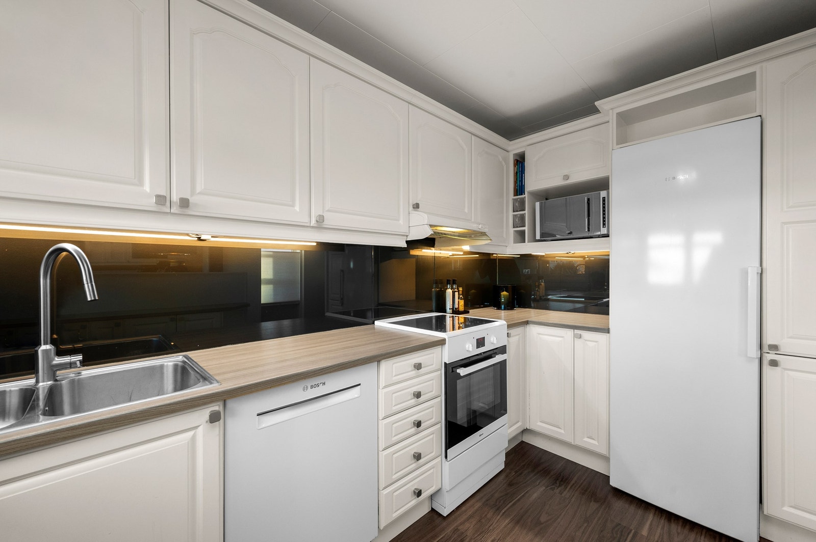 Videre er kjøkkenet utstyrt med frittstående komfyr, oppvaskmaskin og kjøleskap og har rikelig med skap- og benkeplass å boltre seg på. Galleribilde