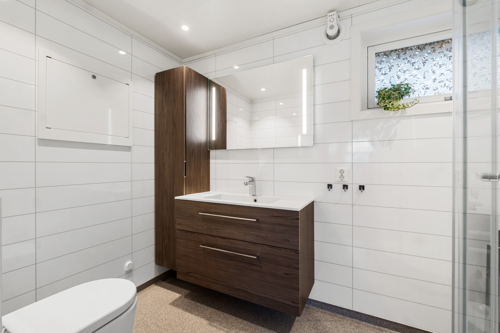 Pent bad med lyse plater på vegg, varme i gulv, mørk innredning, dusjkabinett og wc. Galleribilde