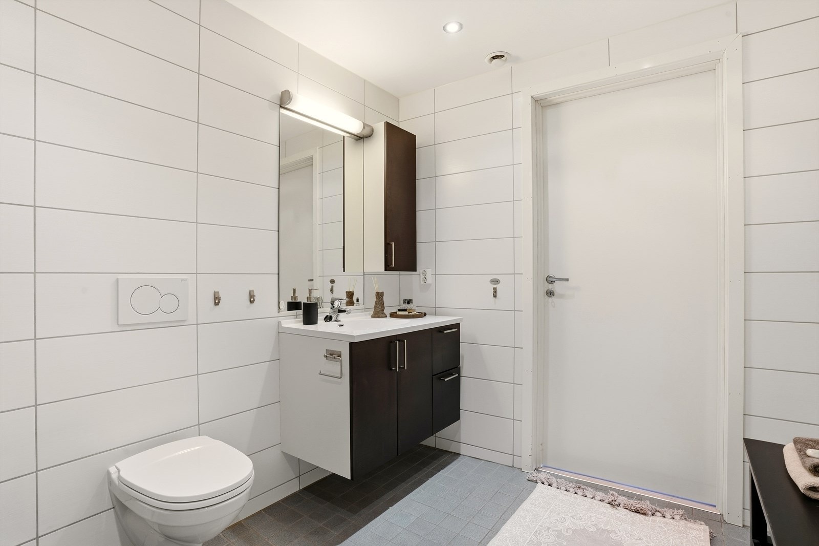 Baderomsinnredningen har en mørk utførelse med servant og rom for oppbevaring. Videre er badet utstyrt med speil, dusjsone med innfellbare dusjvegger og veggmontert toalett. Rommet har opplegg for vaskemaskin. Galleribilde