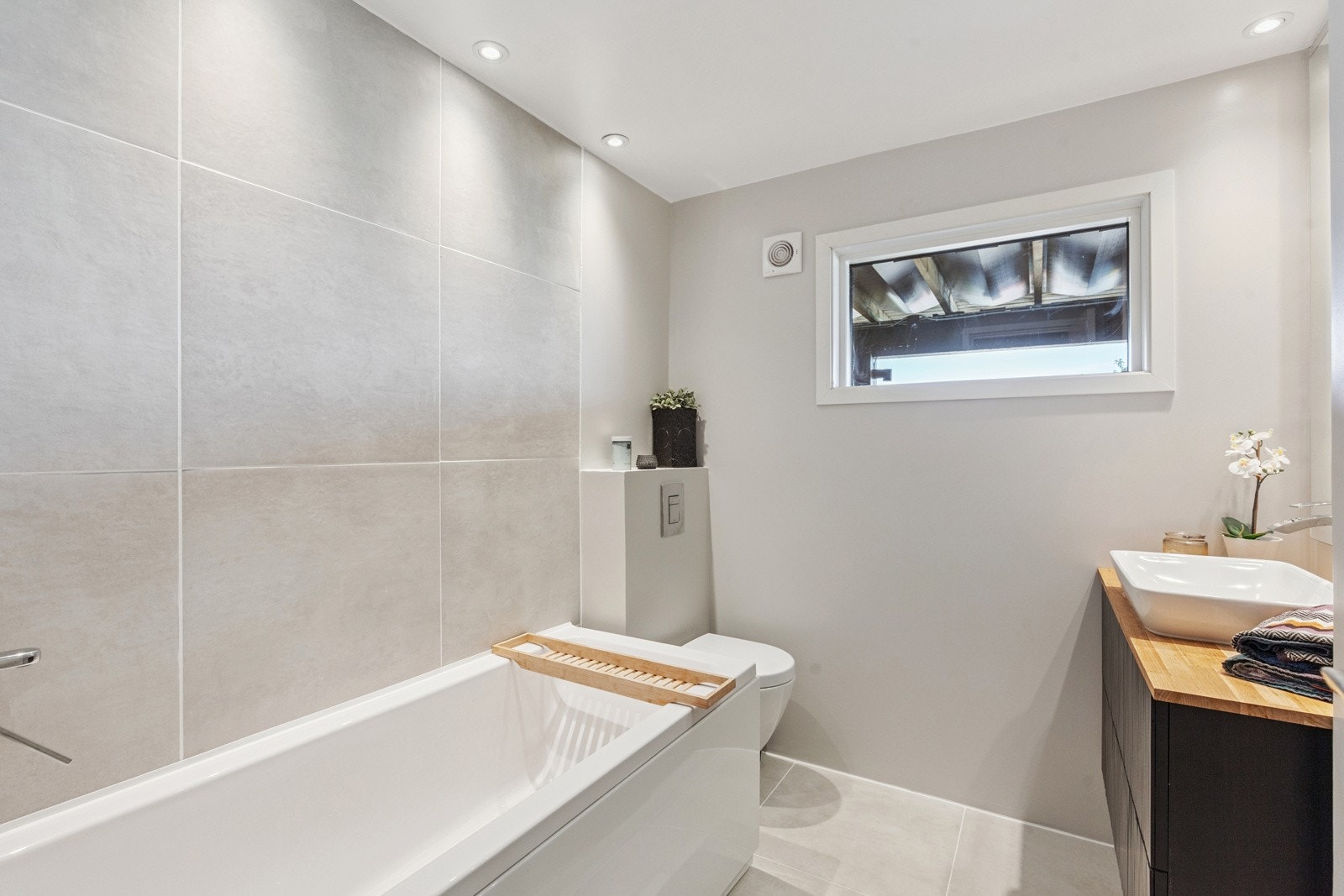 Stilrent bad med flislagt gulv, vegghengt toalett, servant med oppbevaring og badekar Galleribilde