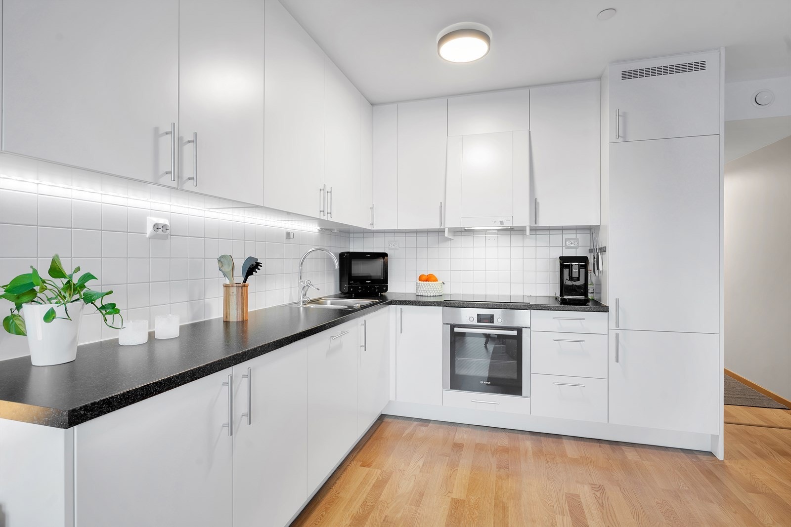 Lekkert kjøkken i hvit utførelse med fliser på vegg over benkeplate. Rikelig med skap- og benkeplass, samt integrerte hvitevarer. Galleribilde