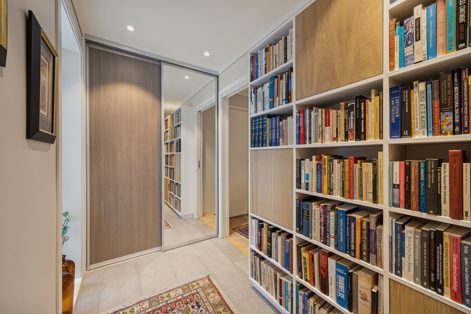 Innbydende entré med stilrene flislagte gulv, praktisk skyvedørsgarderobe og plassbygd bokhylle med integrert skapløsning. Galleribilde