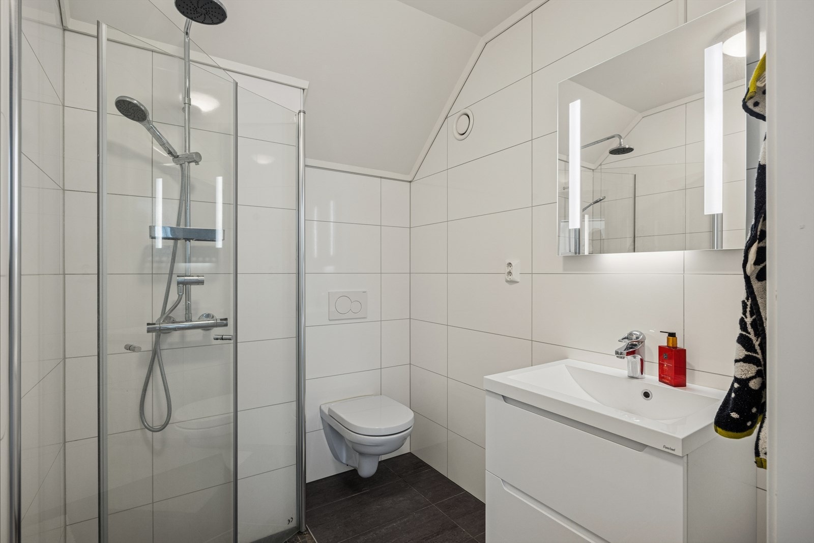 Lyst og flislagt bad med nedfelt servant, veggmontert toalett og dusjhjørne med innfellbare dører. Galleribilde