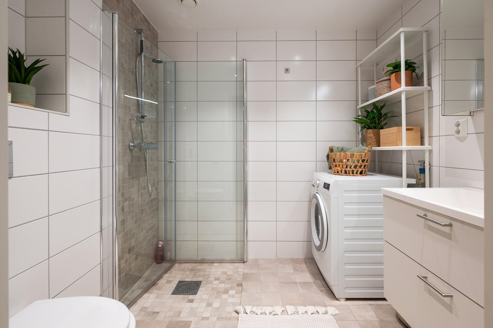 Delikat, romslig baderom med hvite veggfliser, beigemelerte fliser på gulv med varmekabler, samt downlights i himling og opplegg for vaskemaskin Galleribilde