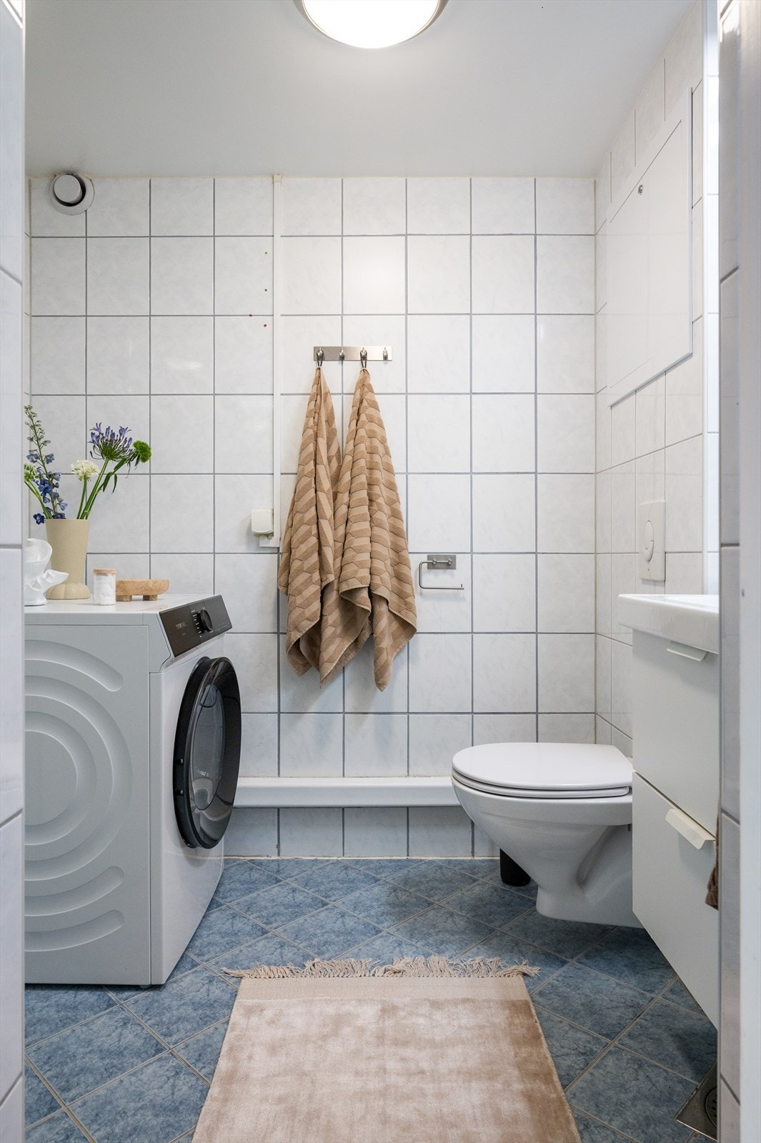 Flislagt baderom innredet med vegghengt toalett, servant med tilhørende innredning og speilskap med belysning. Galleribilde