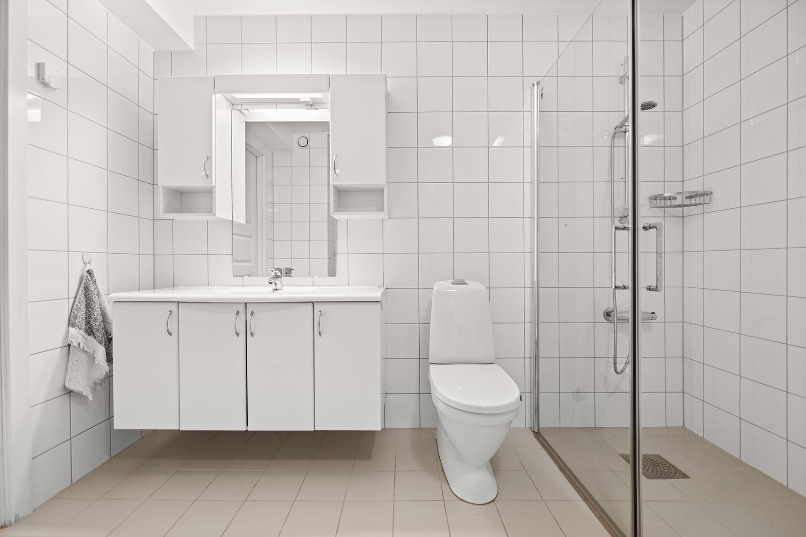 Badet har lyse fliser, dusjkabinett, toalett og en stor baderomsinnredning med speil og skap. Galleribilde