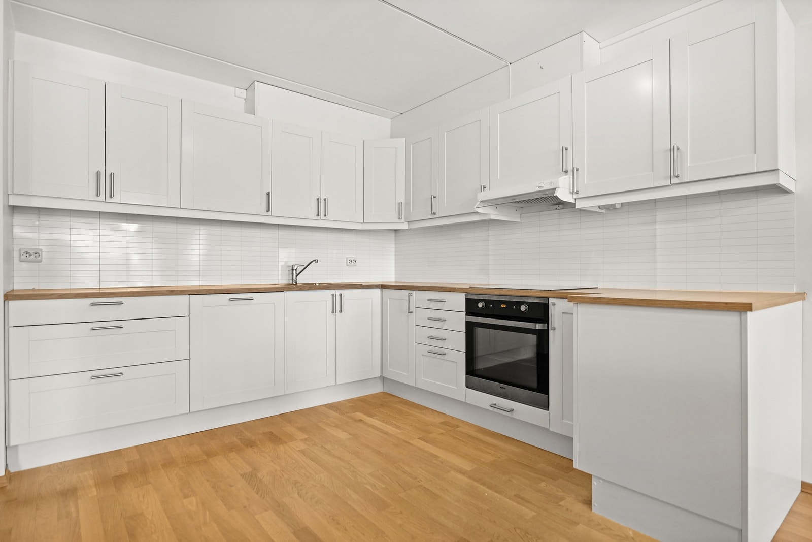 Lyst kjøkken med hvite skap, innebygd komfyr og ventilator. Flislagt vegg over benkeplate. Galleribilde