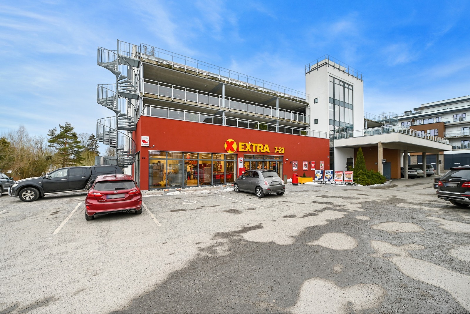 Dagligvarebutikk med parkeringsplasser foran og etasjer over. Butikken har åpningstider fra 7 til 23. Galleribilde