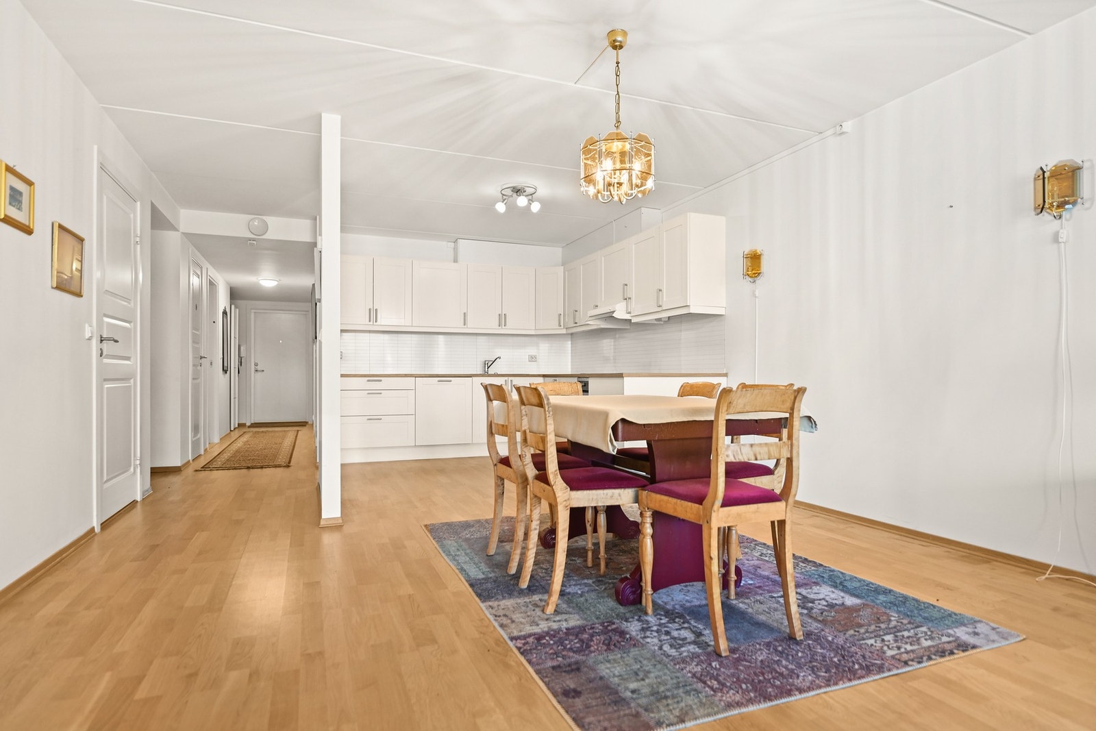 Lyst kjøkken med åpen løsning mot spiseplass og gang. Veggene er malt i hvitt, og gulvet er i tre. Galleribilde