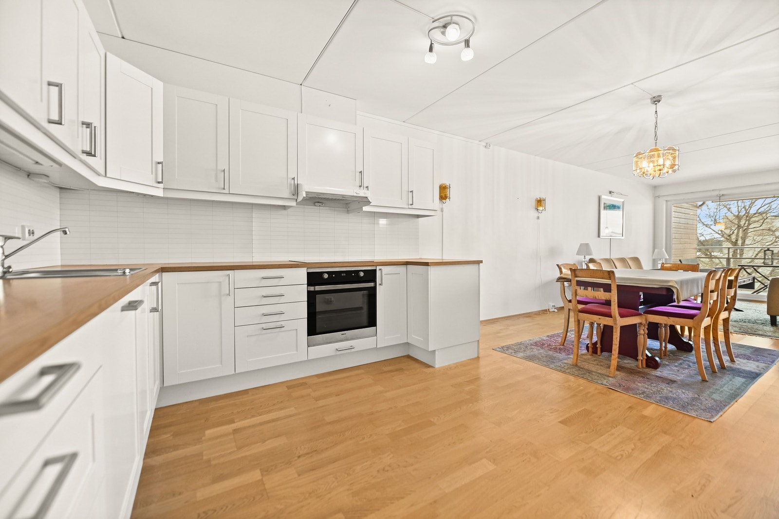 Kjøkken med hvite skap, trebenker og integrerte hvitevarer. Spiseplass med lyse vegger og stor vindusflate. Galleribilde