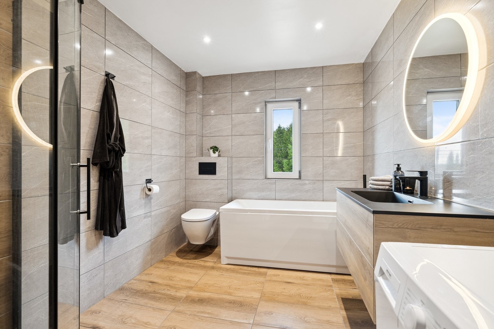 Dusjhjørne, badekar med dusj, vegghengt toalett og servantskap med servant og speil med lys Galleribilde