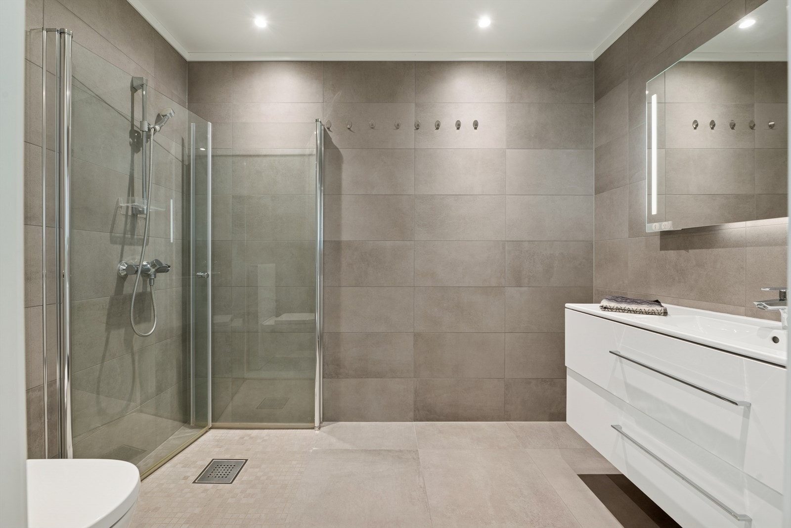 Badet i u. etasje er også innredet med en stilren baderomsinnredning, speil med belysning, veggmontert toalett og dusj med innfellbare dusjdører. Galleribilde