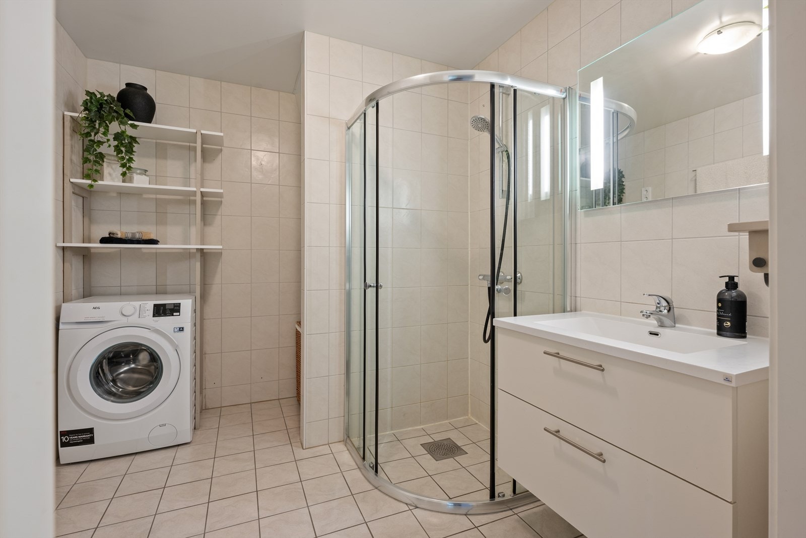 Helfliset baderom med dusjhjørne, vegghengt toalett, opplegg for vaskemaskin, bred servant og speil med belysning Galleribilde