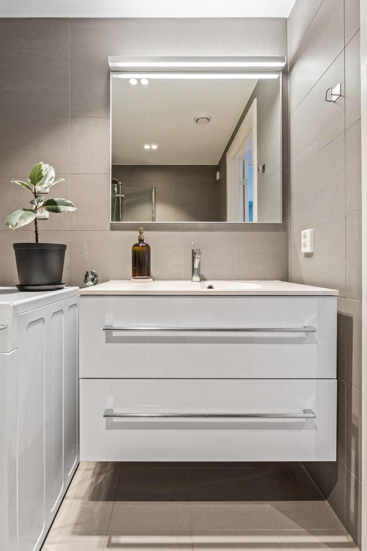 Badet er utstyrt med downlights i himling, vegghengt toalett, dusjhjørne med glassdører, servantinnredning med skuffer og opplegg for vaskemaskin. Galleribilde